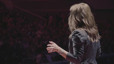 The female financial coach emotional gesturing talks from the stage with spectators at forum. Too many anonymous persons workers and students seat in large auditorium and watch workshop background