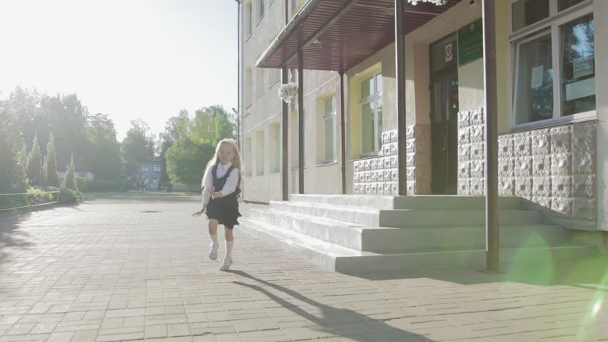 A little funny schoolgirl in school uniform and a briefcase on her shoulders runs out of school. Steadicam shot