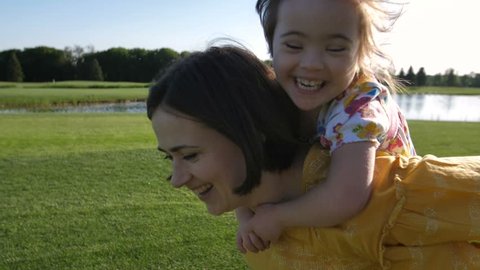 Slow motion of young mother and special needs daughter running on green grass meadow. Mother giving piggyback ride to toddler girl with down syndrome. Delighted girl smiling and laughing in delight