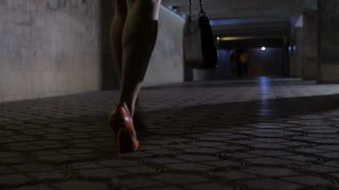 Close-up drunk female legs in high heeled shoes and mini skirt walking alone in dark underpass tunnel at night. Drunken woman stumbling and walking in circles in stiletto shoes