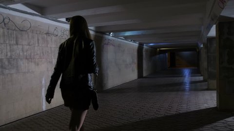 Woman walking alone at night in dark tunnel underpass. Male in hooded jacket robbing female and stealing her bag. Criminal taking the bag and running away to darkness. Steadicam stabilized shot