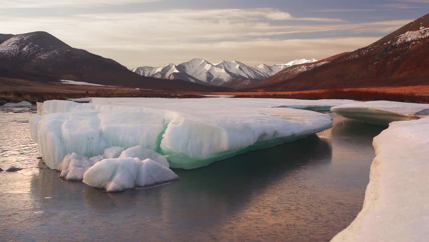 Aerial melting glacier in the river on the background of snow-capped mountain valley.
