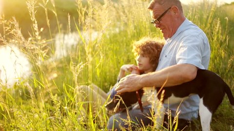 A mature man and a boy are sitting on the river bank. Grandfather is showing something to his grandson hand in the distance. Friendly conversation, male conversation, sunset, happy weekend