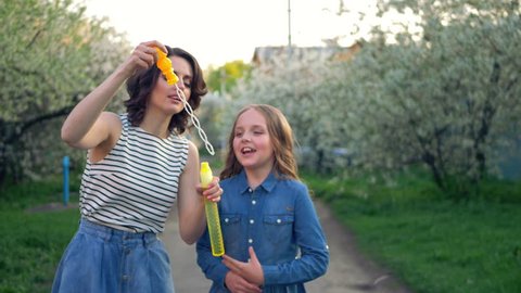 Happy brunette mother and little daughter blow bubbles on the street. They are dressed in stylish blue denim skirts. Beauty Mum and her Child in Park together. Family, Mother's Day Joy. Mom and Baby. 