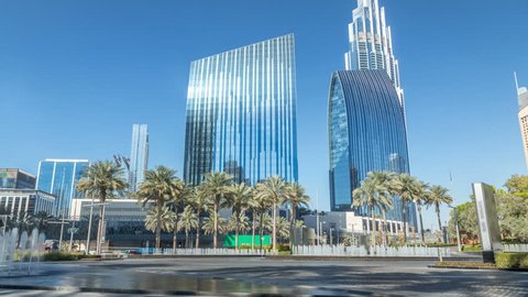 Fountains near main entrance to the tallest skyscraper timelapse, Dubai. Palm and traffic on the road in Dubai downtown at sunny day with modern towers on background