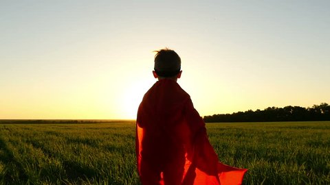 A child in a superhero costume in a red raincoat is running on a green lawn against the backdrop of a sunset simulating a flight, at a slow pace