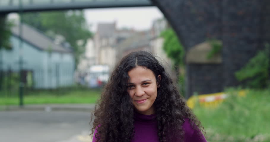 Close up portrait of pretty smiling Young Mixed Race teen Girl with brown curly afro hair, an inner city landscape of a laughing Bi-Racial beautiful student woman against British bridge and housing. 