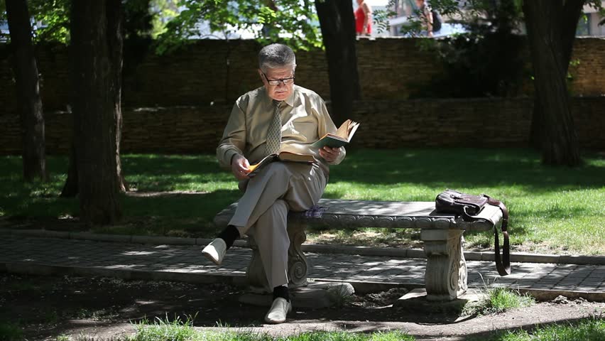 Portrait of a professional old man sitting on a bench and reading two books with a stony parapet behind his back on a sunny day in summer