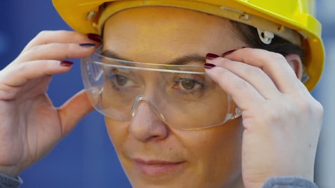 Close up face of middle-aged female engineer in yellow safety helmet taking off transparent protective goggles and looking at camera