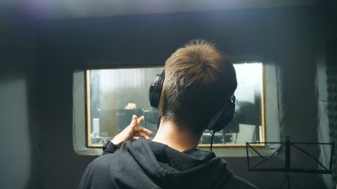 Male singer in headphones singing song at sound studio. Unrecognizable young man emotionally recording song. Working of creative musician. Show business concept. Slow motion Rear back view