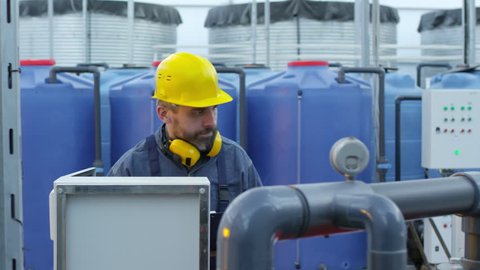 Medium shot of middle aged man with yellow hard hat and earmuffs operating fertigation machine when working in industrial greenhouse complex