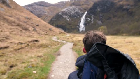 4K Young traveller in Scottish Highlands. Student backpacker nature path trekking through mountains toward waterfall. A natural landscape with a British tourist exploring rural Scotland.