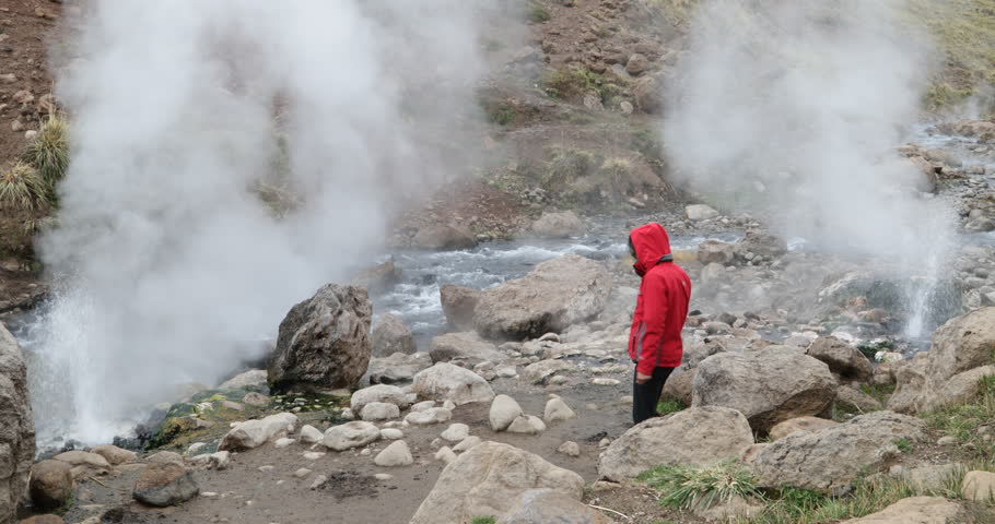 Woman with red jacket walks between blasting geysers, Los Tachos, and climbes rock of Covunco coast warm river. Vapor ascends around the person. Surroundings of Domuyo volcano. Neuquen, Patagonia