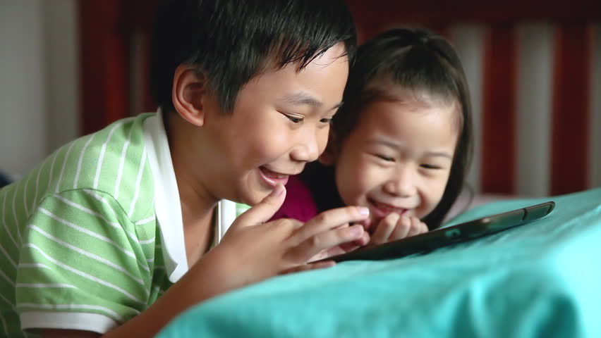Asian children using digital tablet. Happily brother smiling and cheering his younger sister near by. Cute girl playing games excitedly on touchpad and lying prone on bed. Focus shifting from boy to g | Shutterstock HD Video #1012046633