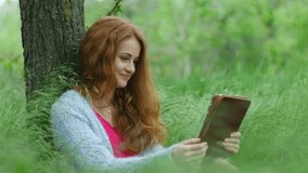 Young, beautiful woman using tablet computer sitting on the grass in the forest. She communicates through a web camera and shows the neighborhood, she looks very cheerful and happy