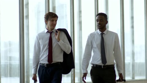 Medium shot of businessmen in suits laughing and chatting while walking along hall of office building with panoramic windows