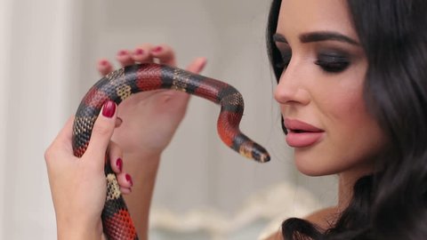 A sexy girl in a dress and with red eyes is holding a snake in her hands. Brunette model with fashion perfect make-up. Snake.
