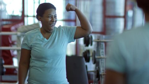 Medium shot of charismatic black woman having fun in gym: she looking in mirror and flexing muscles while laughing and making faces