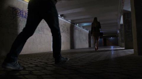 Close-up male attacker legs following young female in mini skirt at night in dark pedestrian underpass. Scared female looking back at criminal and starts running. Steadicam shot