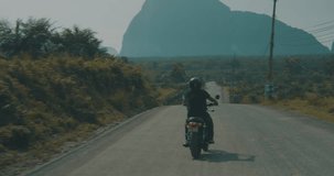 Back view of cool traveler driving his chopper motorbike on tropical island road with beautiful mountain landscape during cloudy early morning - video in slow motion