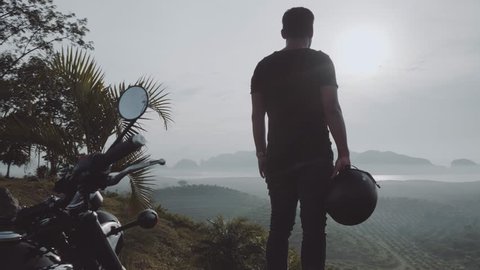 Back view of cool traveler standing in front of his chopper motorbike and admiring beautiful tropical mountain view during cloudy early morning - video in slow motion