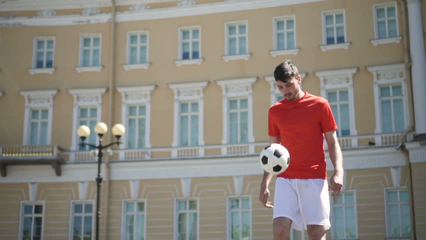 Handsome male football player kicking ball alone by his feet and breast on beautiful city square with antient building on the background on sunny day. Portrait. Outdoors.