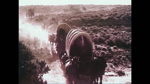 1950s: Train of wagons in desert. Man on horseback. Low angle view of man with gun. People socializing by wagons. View of man. Tracking shot of boy walking next to wagon.