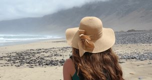 Back view of young woman with straw hat walking on Caleta Famara beach and looking to the ocean, Lanzarote, Spain