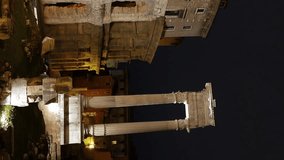 Teatro Di Marcello. The ancient Roman theatre dominates the frame with its monumental travertine and tufa facade, featuring multiple tiers of arched openings framed by engaged Doric and Ionic columns. The structure rises against a deep night sky, its weathered stone surfaces illuminated by warm artificial lighting that accentuates the texture of the masonry and the rhythmic repetition of the arches. Adjacent to the theatre, the remains of the Temple of Apollo Sosianus stand with three reconstructed Corinthian columns visible in the foreground, creating a layered composition of Roman ruins. The camera maintains a steady low-angle perspective, slowly panning upward to reveal the full height of the theatre's curved exterior wall and the later Renaissance palace built atop the ancient structure. The scene is set in the historic Sant'Angelo district of Rome, between the Capitoline Hill and the Tiber River, capturing the monument's integration into the dense urban fabric of the city. The atmosphere is serene and monumental, with the golden glow of the spotlights contrasting against the dark sky to emphasize the enduring presence of this architectural landmark. Originally commissioned by Julius Caesar and completed by Augustus in 13 BC, the theatre was dedicated to the emperor's nephew Marcus Claudius Marcellus and once held up to 20,000 spectators. Vertical video. - Powered by Shutterstock - Get 15% off with code: PIKWIZARD15