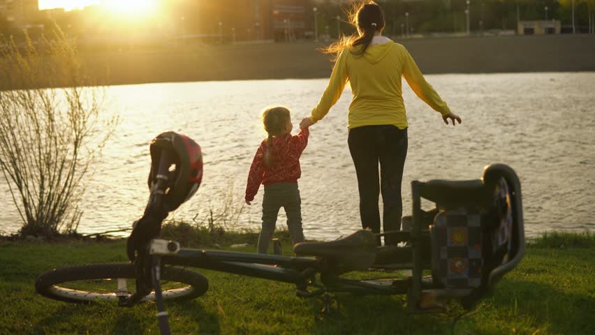 Mom and little daughter enjoy the sun standing on the river bank next to the bike in the summer at sunset.