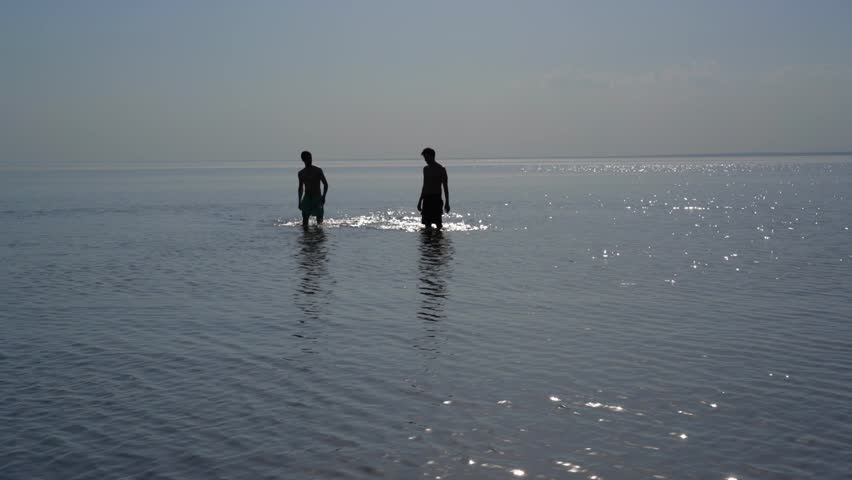 Silhouettes of two young men go knee-deep in the water.