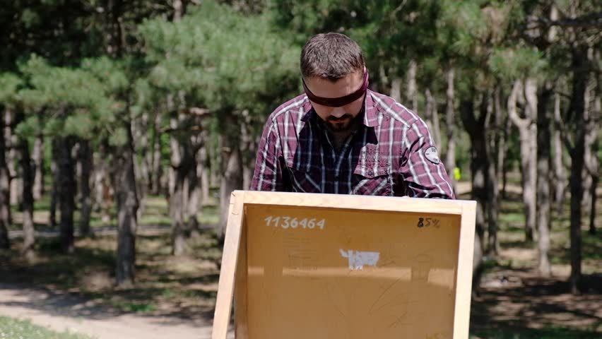 A blondfolded man enthusiastically makes sketches on a canvas while being in the woods