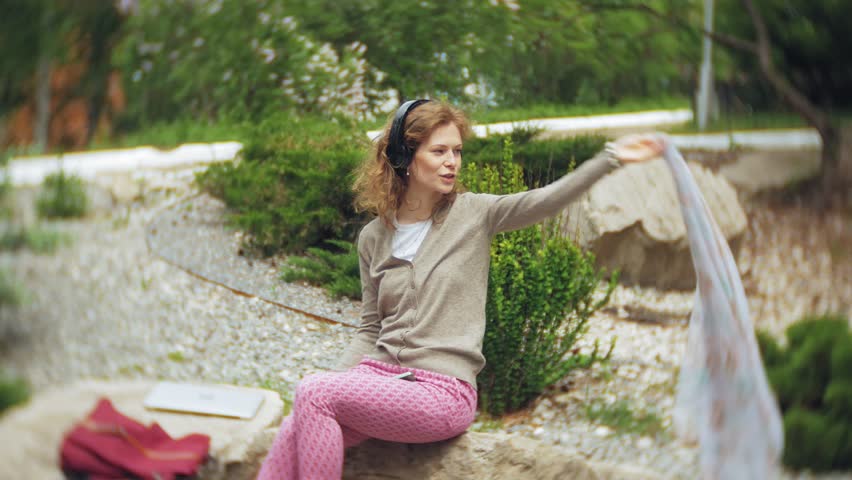 A woman listens to music on headphones, relaxes on the rocks in a beautiful green park. A young perennial woman in the arboretum listens to music. Technology in the open air 4k