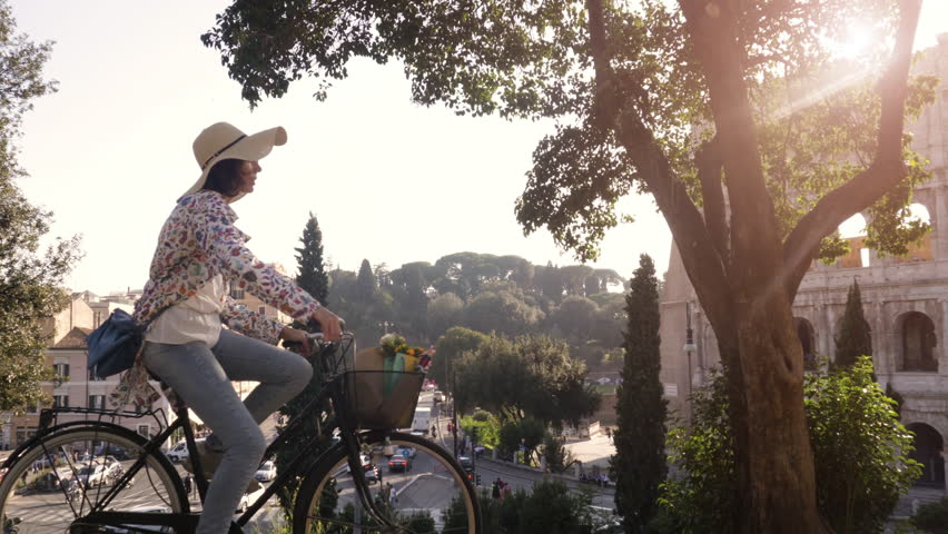 Beautiful young woman in colorful fashion riding bike in front of colosseum in Rome at sunset with trees attractive girl with straw hat steadycam dolly