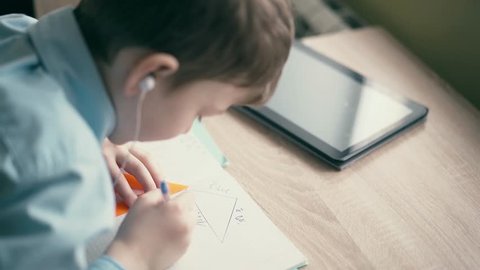 CU: Boy do school homework, writes a ballpoint pen in a notebook. In the ears are inserted headphones, on the table is a tablet computer and school supplies.