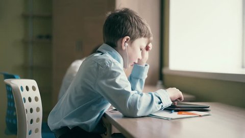 Children do their homework, one sits at the table with notebooks, the second - sits in headphones, closing his eyes and listening to an audiobook. 