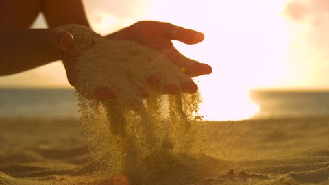 SLOW MOTION, CLOSE UP, LENS FLARE: Unrecognizable female tourist scatters the course sand through her gentle fingertips at beautiful sunset. Cinematic shot of sunrise shining on girl playing with sand