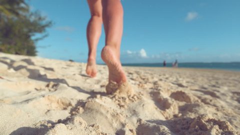 SLOW MOTION, CLOSE UP, LOW ANGLE, DOF: Particles of sand fly in the air as unrecognizable woman jogs barefoot on the tropical beach on remote island. Cheerful girl runs along ocean in the summer sun.