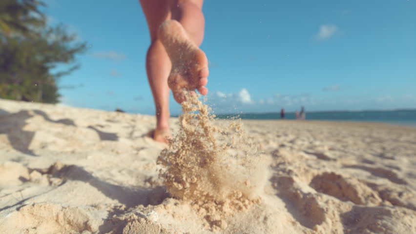SLOW MOTION:, LOW ANGLE, CLOSE UP, DOF Unrecognizable woman running in the hot summer sun along the sunny exotic shore in Cook Islands. Carefree girl takes off her flip flops and runs in the sand. Royalty-Free Stock Footage #1012187819