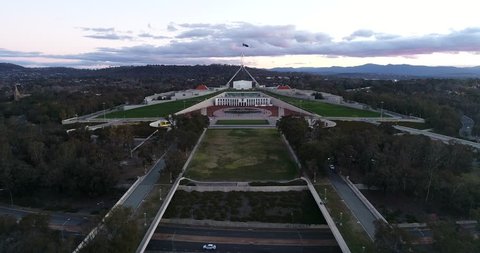 Facade of Australian parliament house on capitol hill in Canberra at sunset time with waving flag on tall flagpole over masts.
