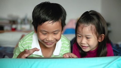 Asian children using digital tablet. Happily sister smiling and cheering her brother near by. Cute boy playing games excitedly on touchpad and lying prone on bed. 