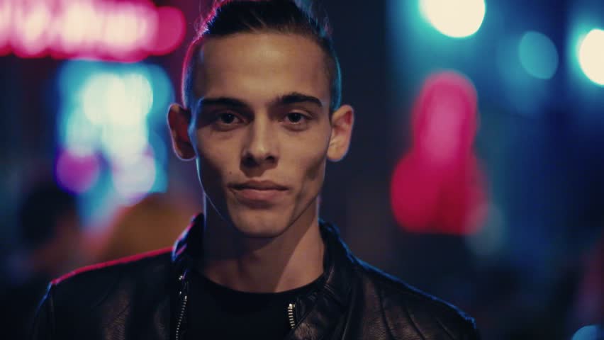 Attractive young man in a leather jacket happily smiles straight to camera. Positive mood, cheerful emotions, feeling good. Male portrait, city lights on the background