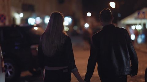 Back view of young people in love holding their hands while walking cross the night city street. Going to the party, club, concert together. Happy together, having fun. Love story