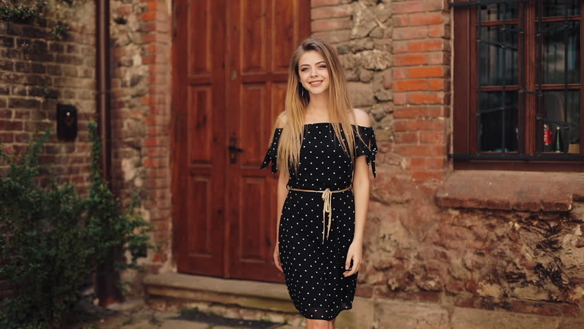 Beautiful girl in black dress walking along the street street old city. Sunny day summer sunshine woman smiling pretty lovely look looking camera smiles glad emotional face leisure. Slow motion