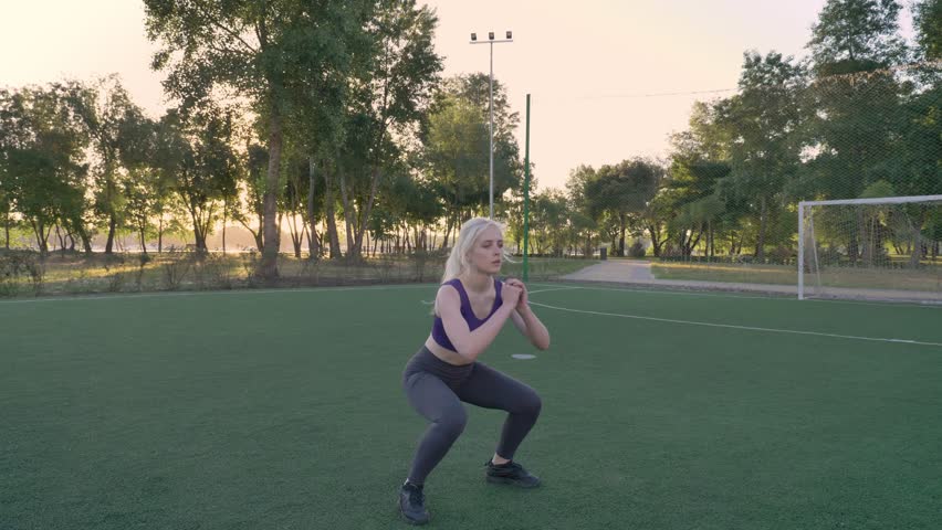 Young blonde woman doing squat jumps exercise on soccer field in park, morning work-out
