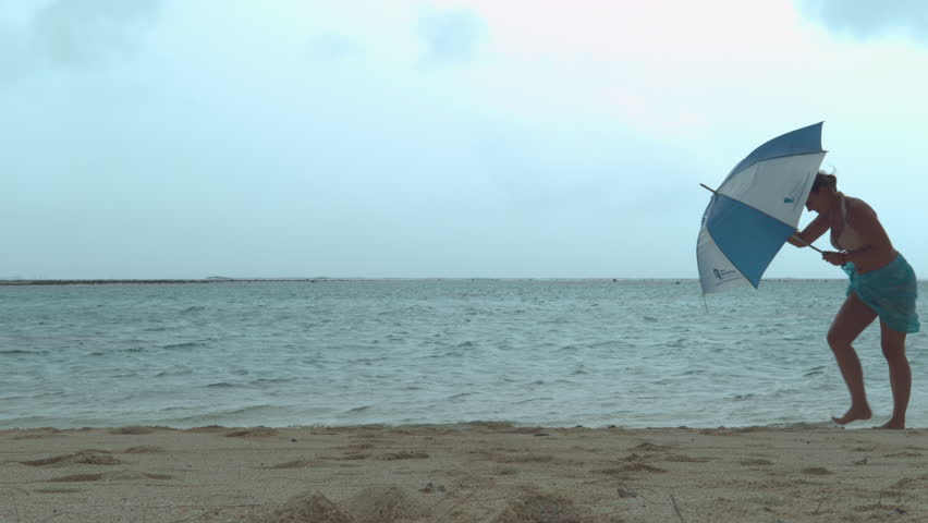 Young tourist struggles to hold her umbrella as she gets caught in a violent windstorm during her relaxing walk along the ocean. Funny shot of woman's umbrella getting destroyed by the powerful wind. Royalty-Free Stock Footage #1012235336