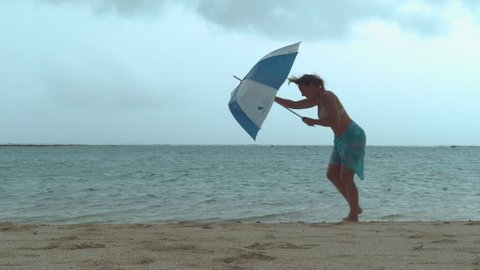Woman's umbrella gets broken by the strong monsoon wind while she walks down the sandy tropical beach. Young female traveler gets caught in horribly windy weather during her trip to the seaside.