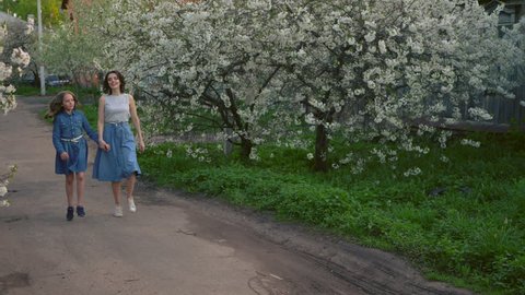 Happy mother with her cute little daughter running down the street holding hands. Brightly blooming trees surround them. Concept mom and daughter together forever, family, blue jeans, love.