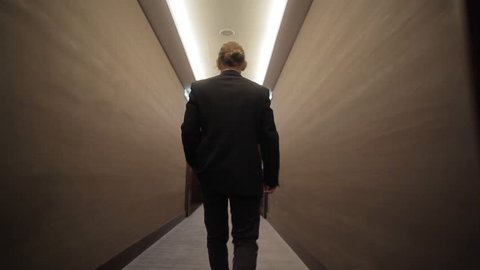 Young businessman going along long narrow corridor with closed doors back view pov camera sliding. Man in business suit clothing walks room hall to meeting work indoors. Office style building success