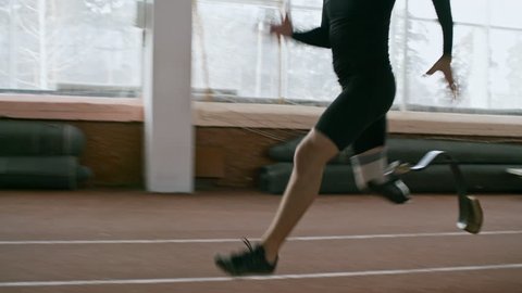 Tracking shot with tilt up of disabled sportsman with artificial fitness leg sprinting on indoor track